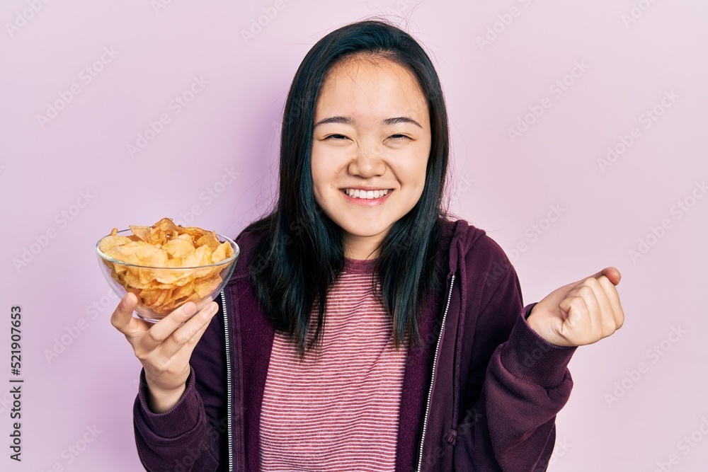 Young chinese girl holding potato chip screaming proud, celebrating victory and success very excited with raised arm Young chinese girl holding potato chip screaming proud, celebrating victory and success very excited with raised arm