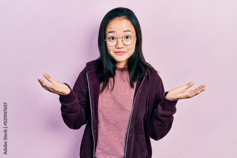 Young chinese girl wearing casual clothes and glasses clueless and confused expression with arms and hands raised. doubt concept. Young chinese girl wearing casual clothes and glasses clueless and confused expression with arms and hands raised. doubt concept.