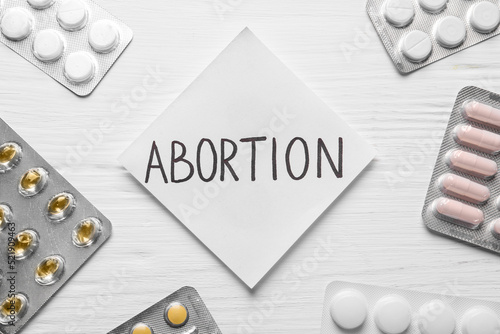 Vászonkép Paper note with word Abortion and pills on white wooden background, flat lay