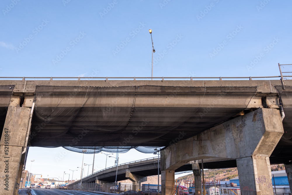 A concrete bridge over a two lane highway. The high overpass has a metal handrail. Black construction netting hangs from under the bridge to catch any concrete debris during construction repairs. A concrete bridge over a two lane highway. The high overpass has a metal handrail. Black construction netting hangs from under the bridge to catch any concrete debris during construction repairs.