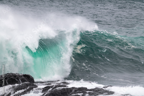 An angry turquoise green color massive rip curl of a wave as it barrels rolls along the ocean. The white mist and froth from the wave are foamy and fluffy. The ocean in the background is deep blue. 