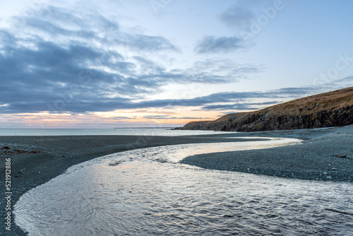 A curved shaped river leading to the Atlantic ocean under a dark blue sky at sunset. The horizon is orange and the glow is on the sand and water. There's a coastline overlooking the sandy beach.