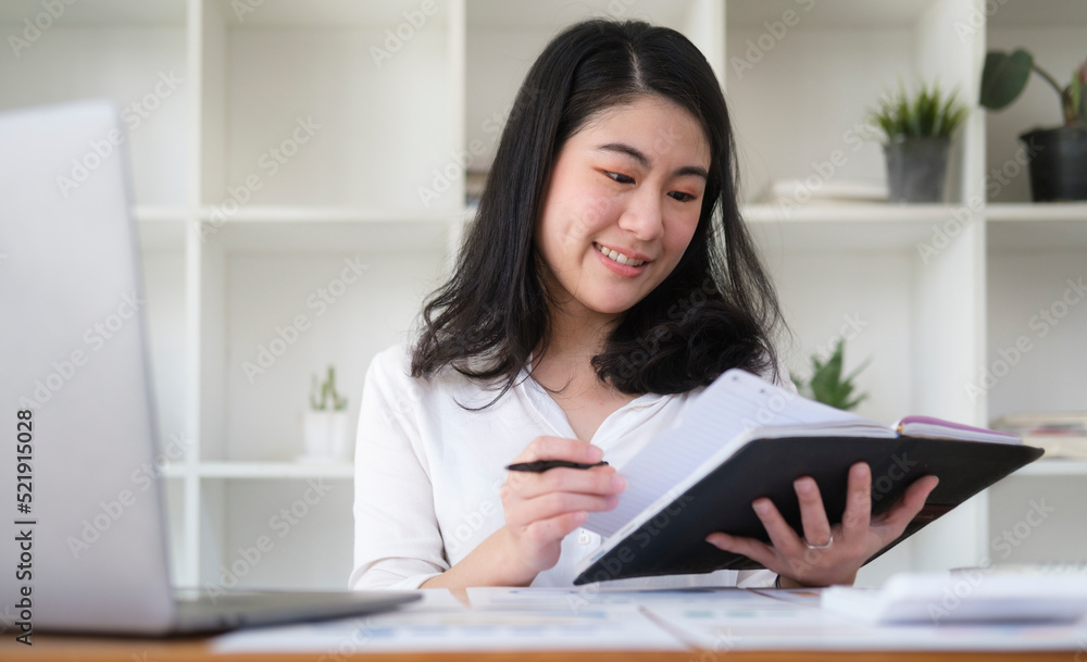 Charming asian businesswoman sitting front of laptop and reading information on notebook. Charming asian businesswoman sitting front of laptop and reading information on notebook.