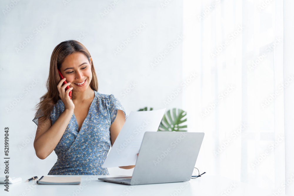 Young pretty Asian business woman talking on the phone when using laptop at working desk in modern office workplace. Young pretty Asian business woman talking on the phone when using laptop at working desk in modern office workplace.
