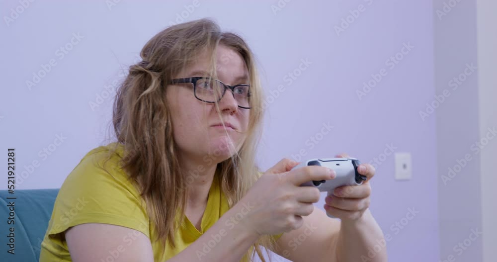 Disheveled, sloppy young woman with crazy look is playing video game using controller for console, hair is getting into face and mouth, but she can not distract herself and tries to blow them away Disheveled, sloppy young woman with crazy look is playing video game using controller for console, hair is getting into face and mouth, but she can not distract herself and tries to blow them away