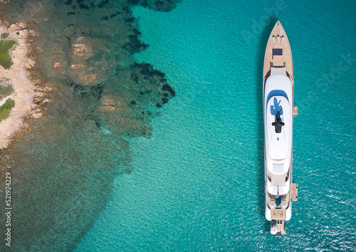 White big yacht for a billionaire anchored off a rocky shore aerial view. Mega yacht on blue water aerial view. Modern big white yacht on transparent water, coastline top view. © Berg