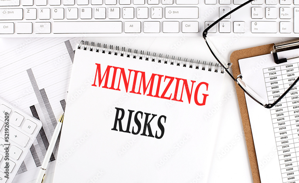 Text MINIMIZING RISKS on Office desk table with keyboard, notepad and analysis chart on white background. Text MINIMIZING RISKS on Office desk table with keyboard, notepad and analysis chart on white background.