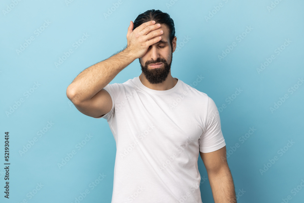 Portrait of unlucky man with beard wearing white T-shirt standing with facepalm gesture, feeling regret and sorrow, blaming herself for mistake. Indoor studio shot isolated on blue background. Portrait of unlucky man with beard wearing white T-shirt standing with facepalm gesture, feeling regret and sorrow, blaming herself for mistake. Indoor studio shot isolated on blue background.