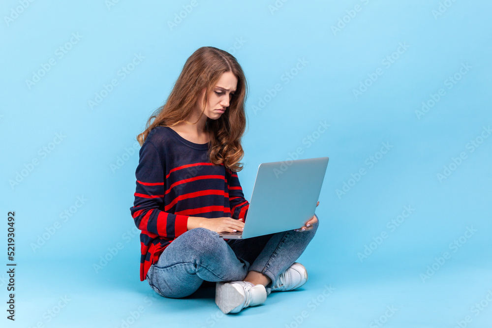 Naklejka premium Portrait of upset woman wearing striped casual style sweater, sitting holding notebook on knees, looking at screen, feeling sadness. Indoor studio shot isolated on blue background.