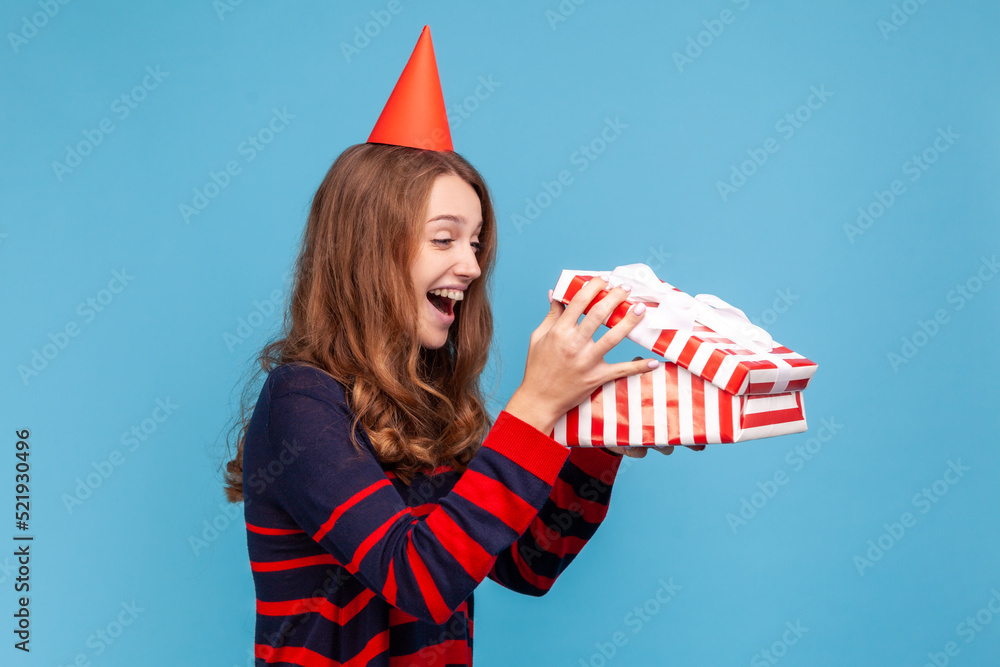 Side view of happy amazed woman in striped casual style sweater and party cone holding present box, looking inside, getting long awaited pleasant gift. Indoor studio shot isolated on blue background. Side view of happy amazed woman in striped casual style sweater and party cone holding present box, looking inside, getting long awaited pleasant gift. Indoor studio shot isolated on blue background.