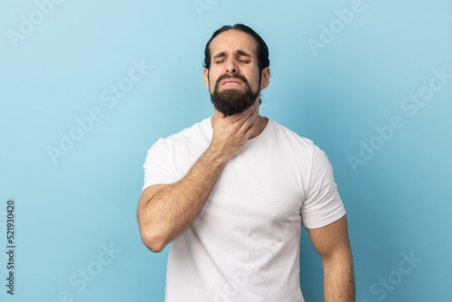 Portrait of unhealthy man with beard wearing white T-shirt suffering from sore throat, keeping hand on her neck, frowning face and closed eyes. Indoor studio shot isolated on blue background.