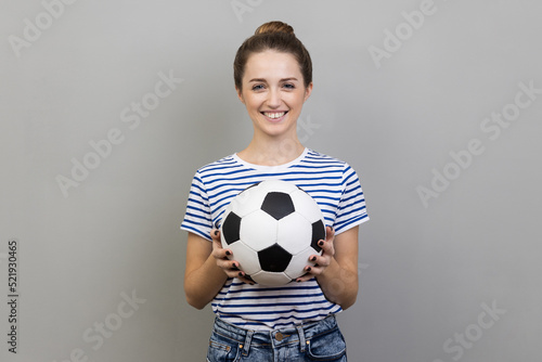 Portrait of woman wearing striped T-shirt looking at camera with pleasant smile, holding soccer ball, watching football match. Indoor studio shot isolated on gray background. © khosrork