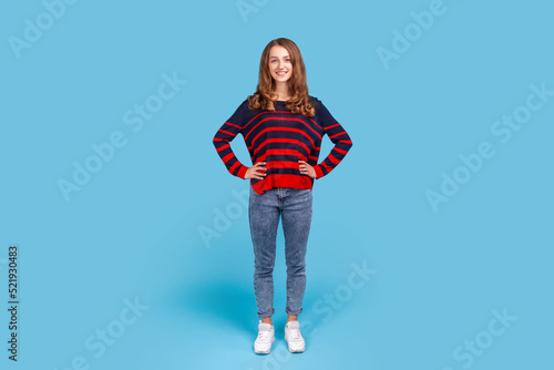 Full length portrait of positive happy woman wearing striped casual style sweater and jeans standing with hands on hips, looking at camera. Indoor studio shot isolated on blue background.