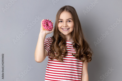 Portrait of little girl wearing striped T-shirt standing holding doughnut and smiling at camera, showing sweet sugary confectionary. Indoor studio shot isolated on gray background.