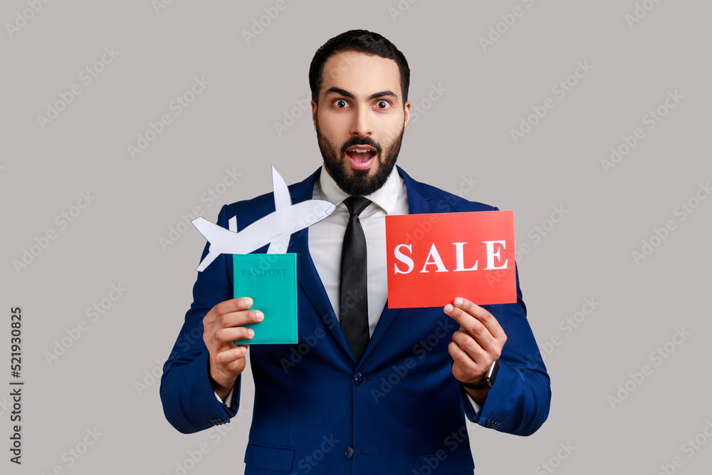 Surprised bearded man holding passport with paper air plane and sale word sign, expressing astonishment, traveling, wearing official style suit. Indoor studio shot isolated on gray background. Surprised bearded man holding passport with paper air plane and sale word sign, expressing astonishment, traveling, wearing official style suit. Indoor studio shot isolated on gray background.