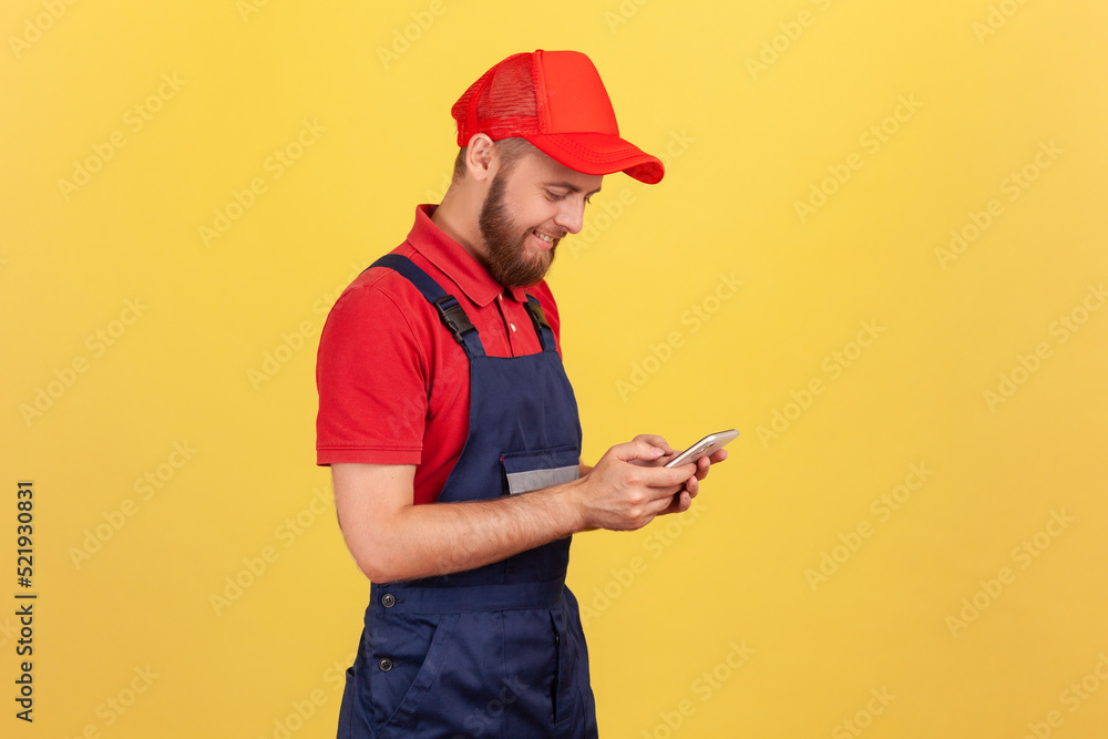 Side view of smiling worker man typing on mobile phone, using cellphone messenger to accept online order as delivery, repair and maintenance services. Indoor studio shot isolated on yellow background. Side view of smiling worker man typing on mobile phone, using cellphone messenger to accept online order as delivery, repair and maintenance services. Indoor studio shot isolated on yellow background.