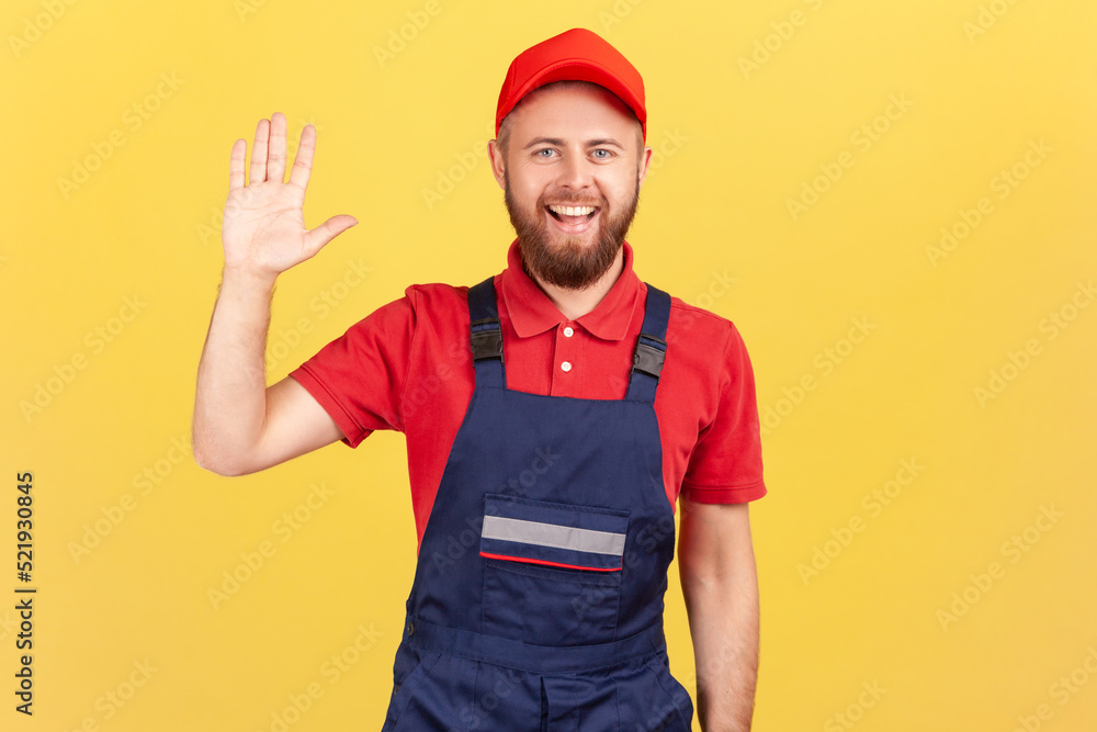 Portrait of delighted worker man standing and looking at camera, waving hand, saying hello or goodbye, wearing blue overalls and red cap. Indoor studio shot isolated on yellow background.