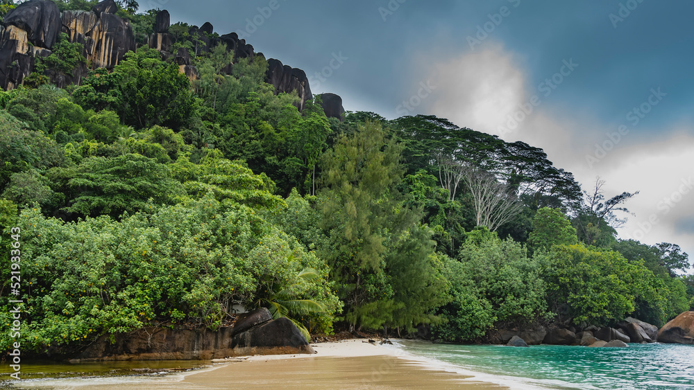 Sandy beach on a tropical island. The rocky slopes of the mountain are overgrown with lush vegetation. The turquoise ocean is calm. Clouds in the blue sky. Seychelles. Mahe
