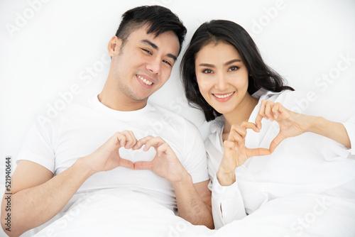 Asian lover show heart sign with hand, they feeling happy and smiling, they rest on bed, happiness honeymoon