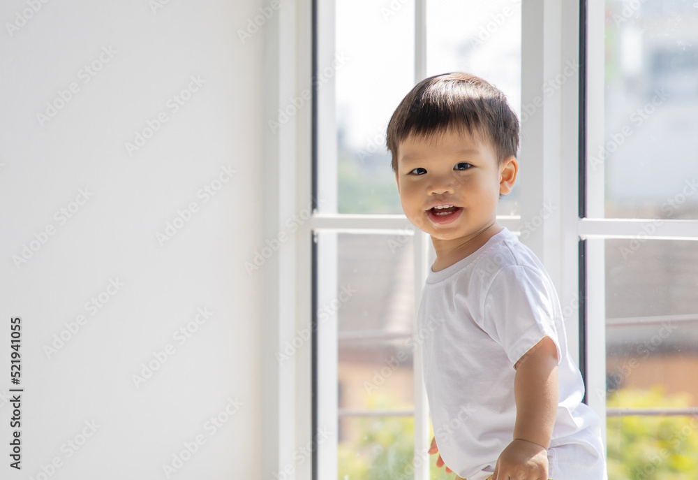 Little asian boy standing and smiles in front of big window at home. Little asian boy standing and smiles in front of big window at home.