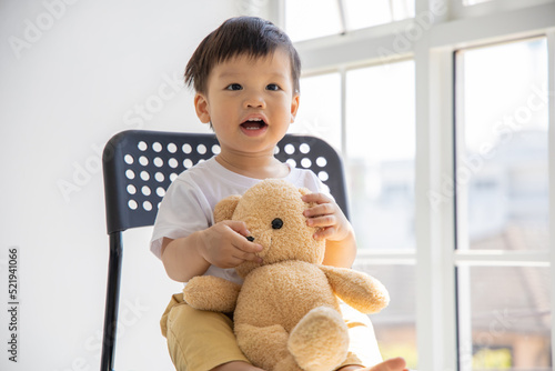 Little asian boy sitting on black chair and holding bear doll with smile in front of big window at home.