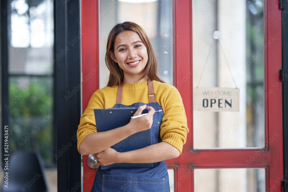 Charming Asian businesswoman in an apron hold an order book in front of a cafe counter with a beaming smile preparing to open and take orders.
