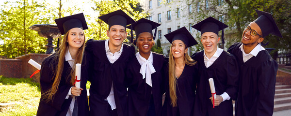 Happy diverse university graduates in green campus yard after graduation. Cheerful joyful students in square academic caps standing together, looking at camera and smiling. Group portrait. Banner
