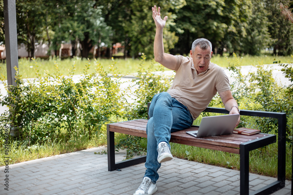 Senior happy and rejoicing gray haired businessman sitting on bench with raised arm on green city park near open laptop. Modern lifestyle, work success, good news. Technology and people concept Senior happy and rejoicing gray haired businessman sitting on bench with raised arm on green city park near open laptop. Modern lifestyle, work success, good news. Technology and people concept