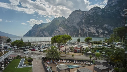 Time lapse of the port area of Riva del Garda and the clouds moving over the lake and the mountains. Transport of vehicles along the lakefront and tourists who enjoy the services of the place.