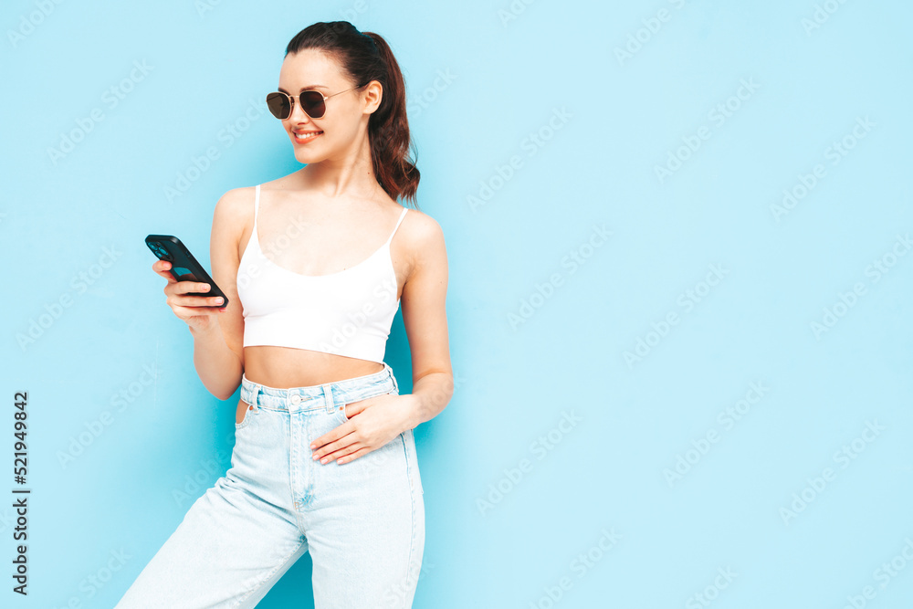 Young beautiful smiling female in trendy summer jeans. Sexy carefree woman posing near blue wall in studio. Positive brunette model looking at cellphone screen. Holding smartphone and using apps Young beautiful smiling female in trendy summer jeans. Sexy carefree woman posing near blue wall in studio. Positive brunette model looking at cellphone screen. Holding smartphone and using apps