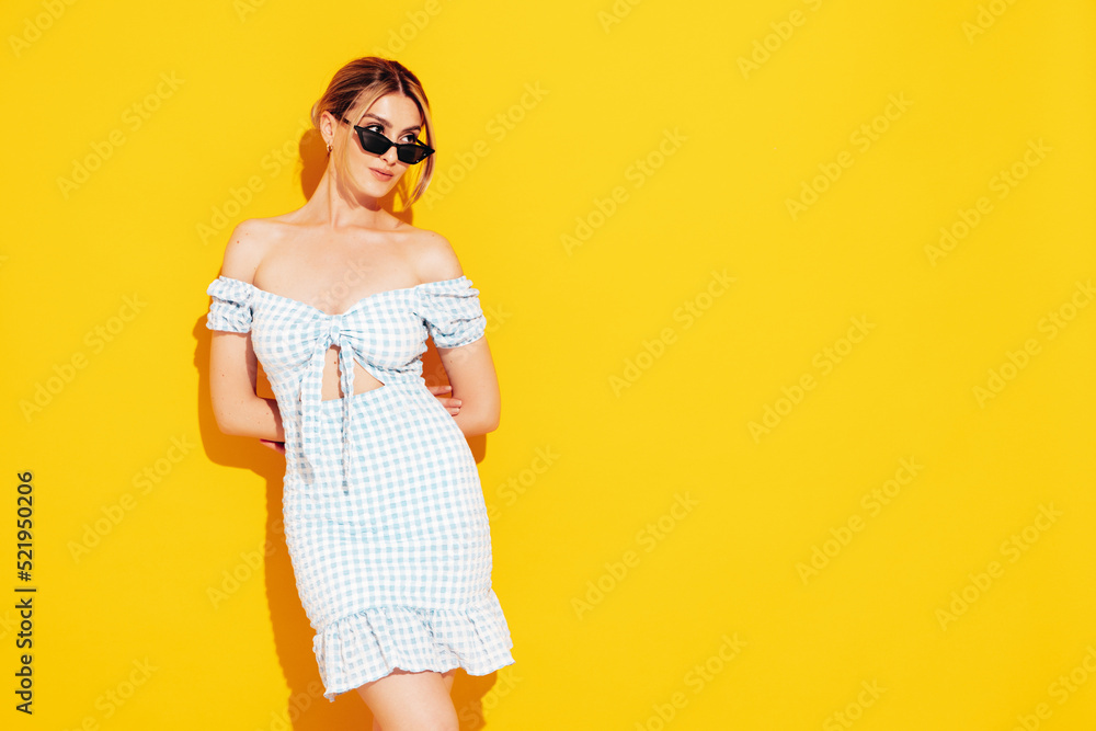 Fototapeta premium Young beautiful smiling female in trendy summer sundress. Sexy carefree woman posing near yellow wall in studio. Positive model having fun. Cheerful and happy. In sunglasses
