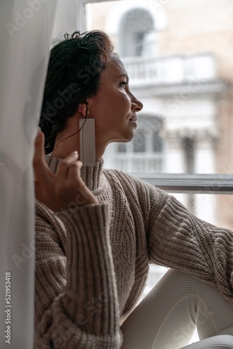 Free time for rest. Profile of a beautiful woman sitting on a white window sill at home, holding a cotton plant in her hands. She is wearing a sweater, leggings and white boots.