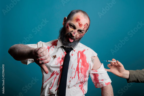 Zombie doing thumbs down sign about vaccine during covid 19 pandemic, looking scary and creepy with bloody wounds. Brain eating apocalyptic devil showing dislike symbol with syringe.