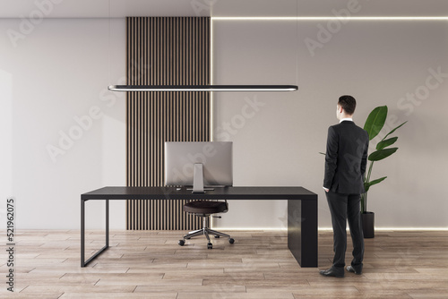 Back view of young european businessman thinking in office interior with furniture, equipment, wooden flooring and concrete wall. CEO, executive and worker concept.