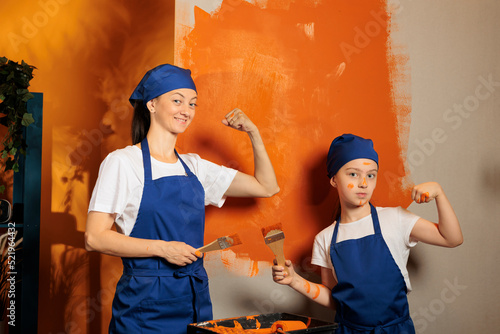 Portrait of mom and kid flexing arm muscles while painting room walls with orange color paint and brush. Woman with little child using renovating tools, working together on house decor.