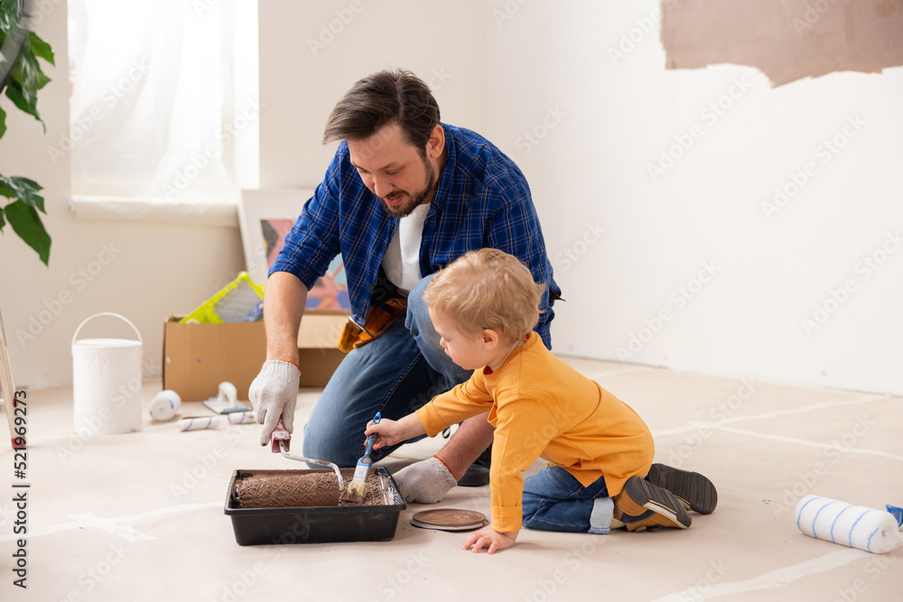 In the room undergoing renovation, a 2-year-old blond boy, together with his brunette dad, put brown paint on a paint roller. The child is very enthusiastic about the process.