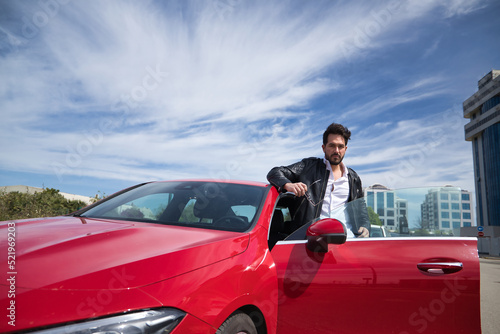 Handsome young man with beard, sunglasses, leather jacket and white shirt, leaning on the roof of his red sports car, looking at the camera seriously. Concept beauty, fashion, luxury, motor, sports. © Manuel