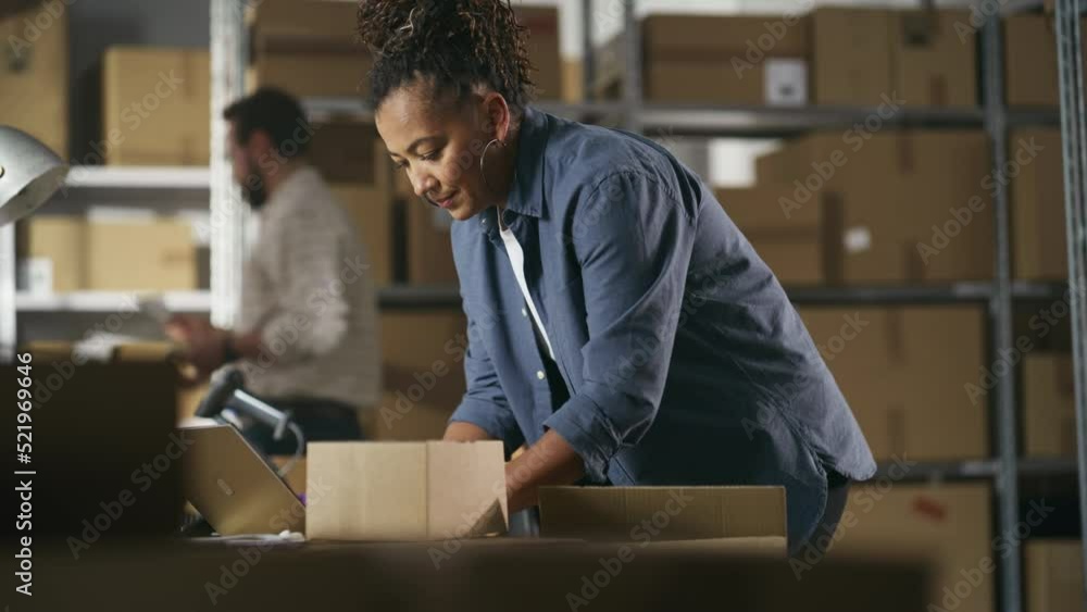Inventory Manager Preparing a Small Cardboard Parcel for Postage. Multiethnic African American Female Small Business Owner Working on Laptop Computer in Warehouse. Handheld Footage. Inventory Manager Preparing a Small Cardboard Parcel for Postage. Multiethnic African American Female Small Business Owner Working on Laptop Computer in Warehouse. Handheld Footage.