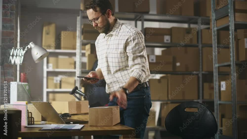 Storeroom Inventory Worker Using Smartphone to Scan a Barcode on Parcel, Preparing a Small Cardboard Box for Postage. Small Business Owner Working on Laptop in Warehouse with Colleague. Storeroom Inventory Worker Using Smartphone to Scan a Barcode on Parcel, Preparing a Small Cardboard Box for Postage. Small Business Owner Working on Laptop in Warehouse with Colleague.