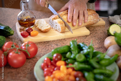 A rustic bread on a wooden cutting board and two woman's hands preparing a snack with a tomato bruschetta and spicy oil. Assortment of fresh vegetables on the table