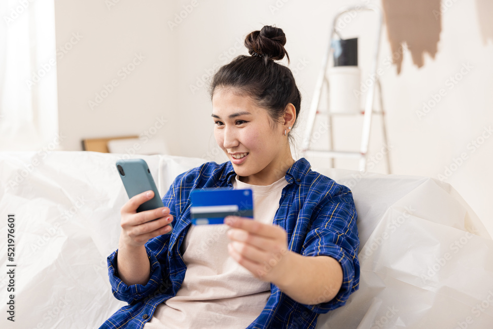 a young and cute girl of Asian appearance, holding a phone in her right hand and a bank card in her left. She makes online purchases for her apartment, which she has started renovating. a young and cute girl of Asian appearance, holding a phone in her right hand and a bank card in her left. She makes online purchases for her apartment, which she has started renovating.