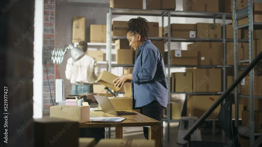 Small Business Owner Works on Laptop Computer while Standing at Her Desk in Warehouse. Employee Working in the Room with Shelves Full of Cardboard Box Parcels in the Background. Small Business Owner Works on Laptop Computer while Standing at Her Desk in Warehouse. Employee Working in the Room with Shelves Full of Cardboard Box Parcels in the Background.