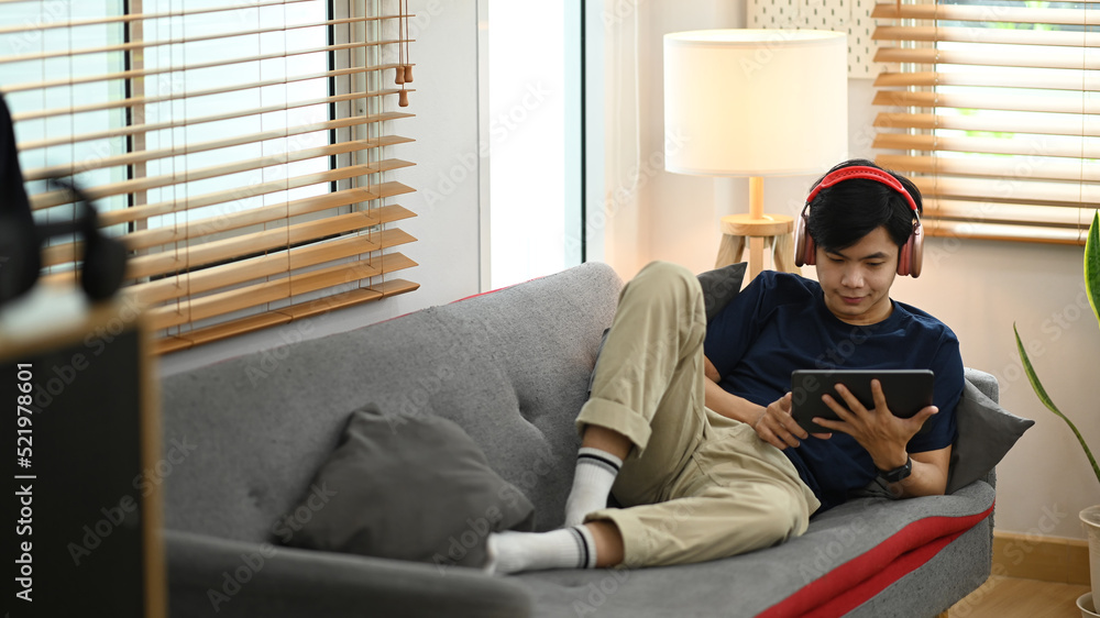 Young asian man using digital tablet, chatting in social networks while lying on couch in cozy living room