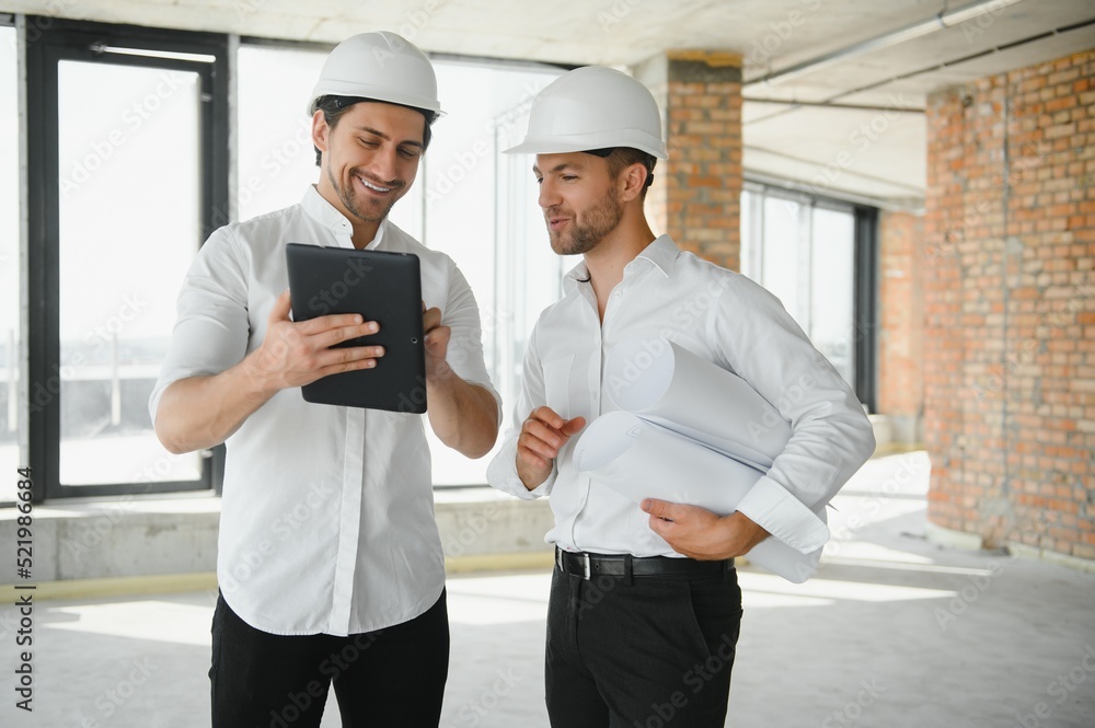 A front view of two smart architects with white helmets reviewing blueprints at a construction site on a bright sunny day