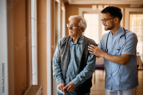 Male caregiver and senior man look through the window ar nursing home.