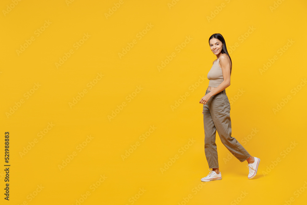 Full body smiling happy fun cool young latin woman 30s she wearing basic beige tank shirt walk stroll going look camera isolated on plain yellow backround studio portrait. People lifestyle concept Full body smiling happy fun cool young latin woman 30s she wearing basic beige tank shirt walk stroll going look camera isolated on plain yellow backround studio portrait. People lifestyle concept