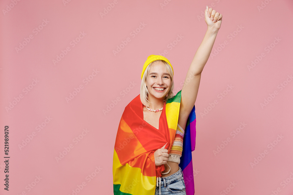 Young confident fun blond lesbian woman she wear yellow hat wrapped in striped flag do hero gesture clench fist isolated on plain pastel light pink background. People lgbtq lifestyle fashion concept. Young confident fun blond lesbian woman she wear yellow hat wrapped in striped flag do hero gesture clench fist isolated on plain pastel light pink background. People lgbtq lifestyle fashion concept.