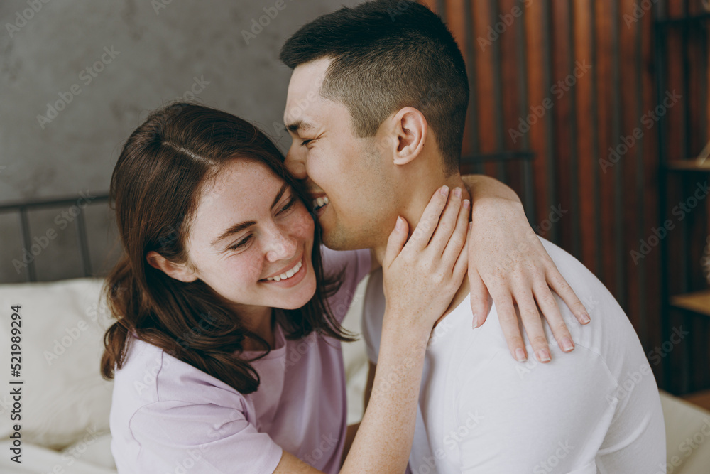 Young smiling happy couple two family man woman she he wear t-shirt pajama hugging cuddling kiss cheek sitting in bed rest spend time together in bedroom home in own room hotel. Real estate concept. Young smiling happy couple two family man woman she he wear t-shirt pajama hugging cuddling kiss cheek sitting in bed rest spend time together in bedroom home in own room hotel. Real estate concept.