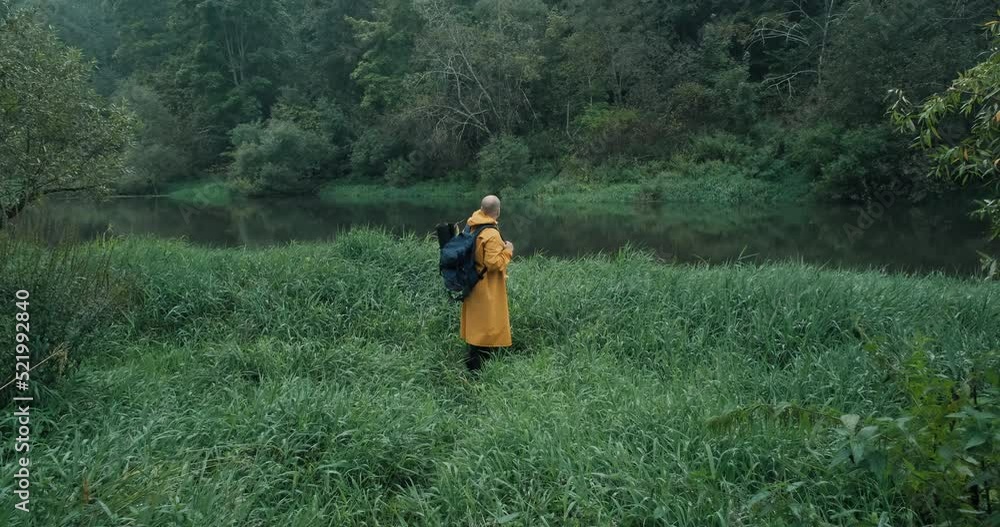 Hiker in a yellow raincoat with a backpack in the tall grass next to the river. The concept of backpacking, lifestyle, hike, extreme hobbies and travel Hiker in a yellow raincoat with a backpack in the tall grass next to the river. The concept of backpacking, lifestyle, hike, extreme hobbies and travel