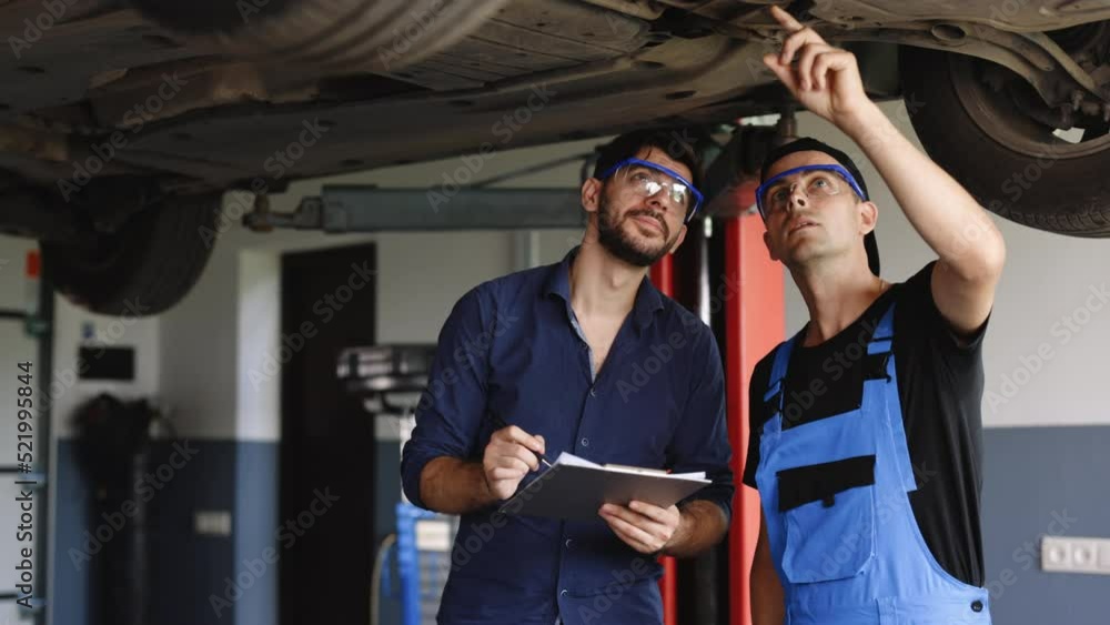 Manager Checks Data on a Notebook and Explains the Breakdown to a Mechanic. Car Service Employees Inspect the Bottom and Skid Plates of the Car. Modern Workshop. Auto Service Manager Checks Data on a Notebook and Explains the Breakdown to a Mechanic. Car Service Employees Inspect the Bottom and Skid Plates of the Car. Modern Workshop. Auto Service